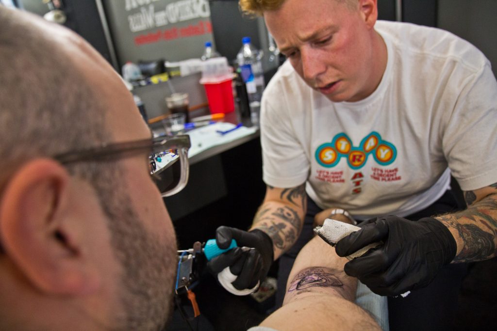 2019 09 13 kpaynter friday the 13th tattoos-8 True Hand tattoo artist Chris Fernandez gives a tattoo of the grim reaper holding a bad news sign to Jamie Santoro. (Kimberly Paynter/WHYY)