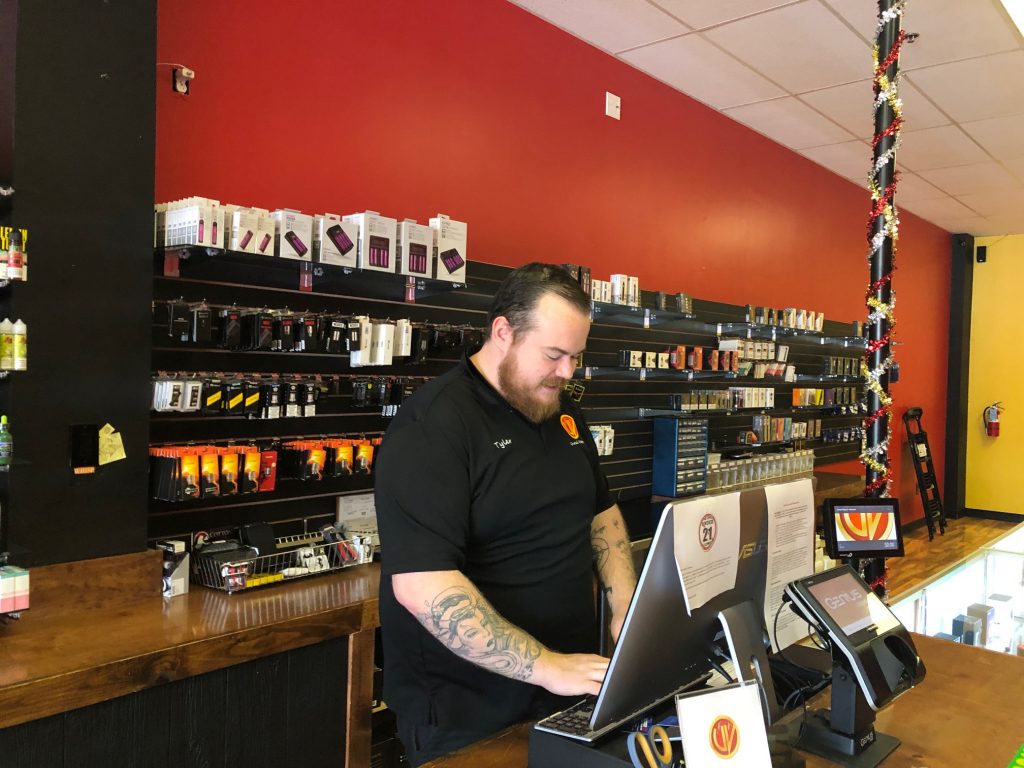 Wilson at register Tyler Wilson, assistant manager at Unreal Vapors near Newport, Del., rings up an order for a lemonade-flavored e-cigarette fluid. (Cris Barrish/WHYY)