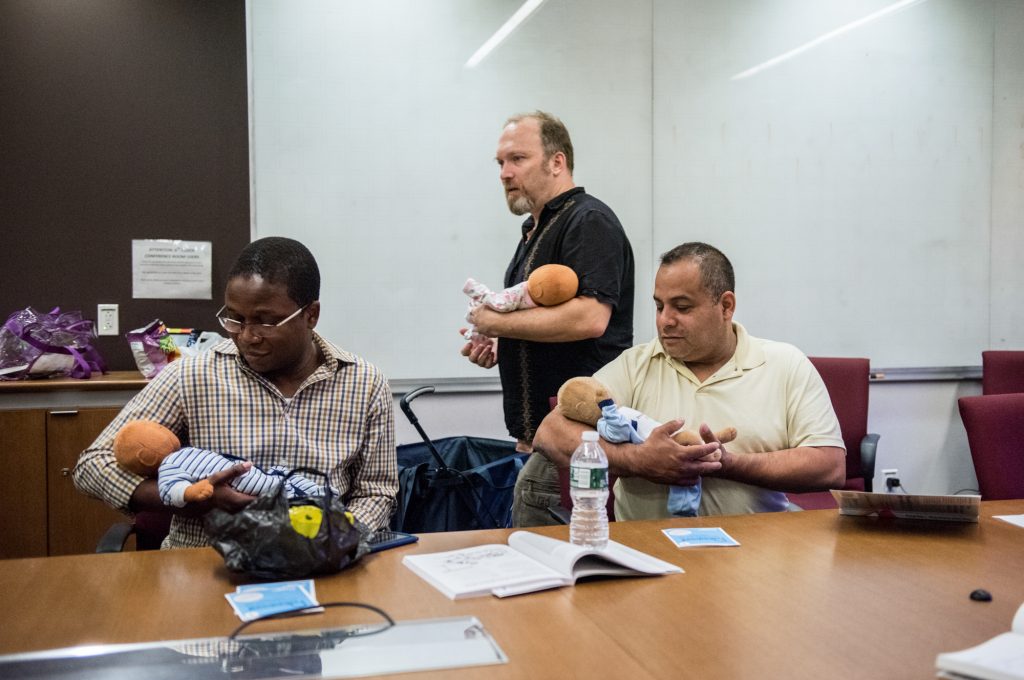 Joe Bay (center), coach of a New York City “Bootcamp for New Dads,” instructs Adewale Oshodi (left) and George Pasco in how to cradle an infant for best soothing. Joe Bay (center), coach of a New York City