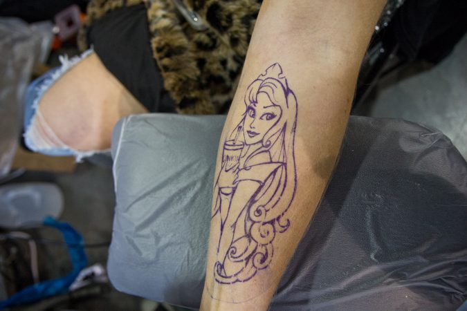 Danny Sun tattoos Disney Princess Aurora drinking Dunkin’ Donuts iced coffee onto Sarah Czechowski. (Kimberly Paynter/WHYY)