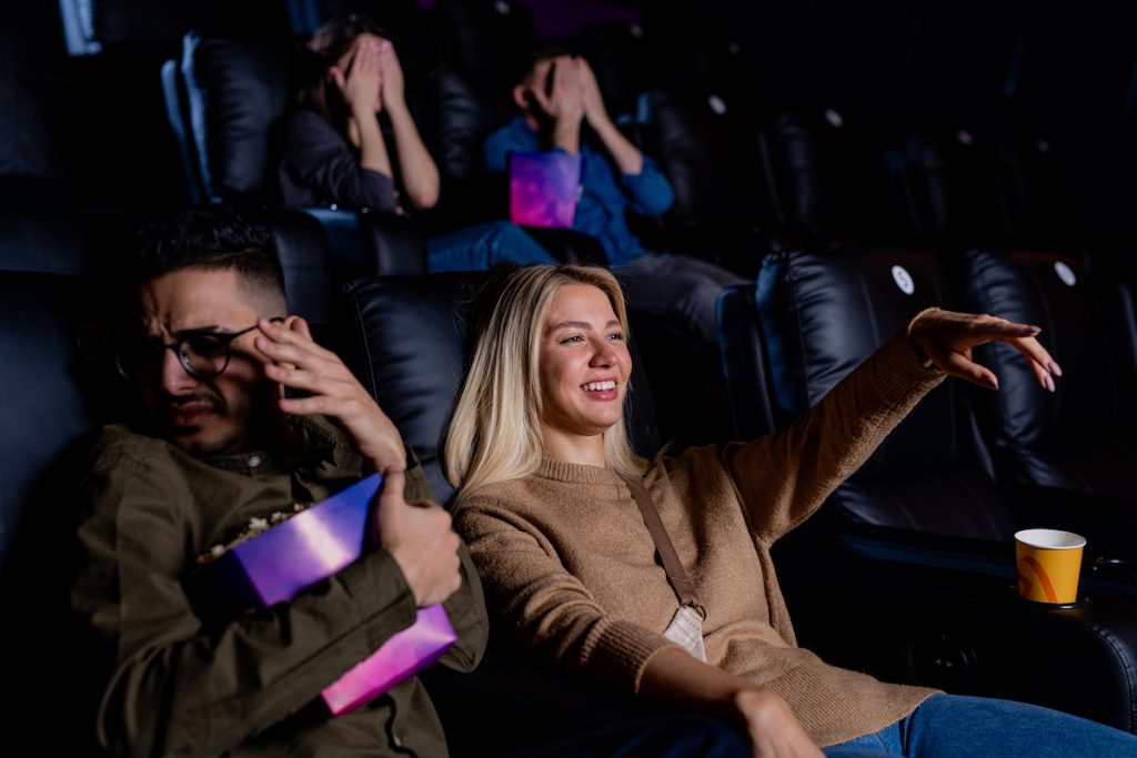 Young smiling woman pointing at large screen while watching action movie with her boyfriend holding Watching movies