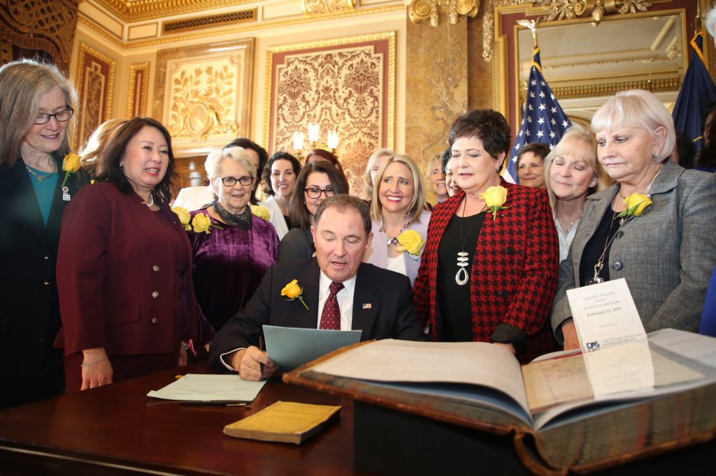 Utah Gov. Gary Herbert signs bill honoring the state’s pioneering women suffragists on Wednesday. He’s surrounded by state senators and representatives, and his wife, who are all wearing the yellow rose symbolizing suffrage. Utah Gov. Gary Herbert signs bill honoring the state's pioneering women suffragists on Wednesday. He's surrounded by state senators and representatives, and his wife, who are all wearing the yellow rose symbolizing suffrage. (Utah Governor's Office)
