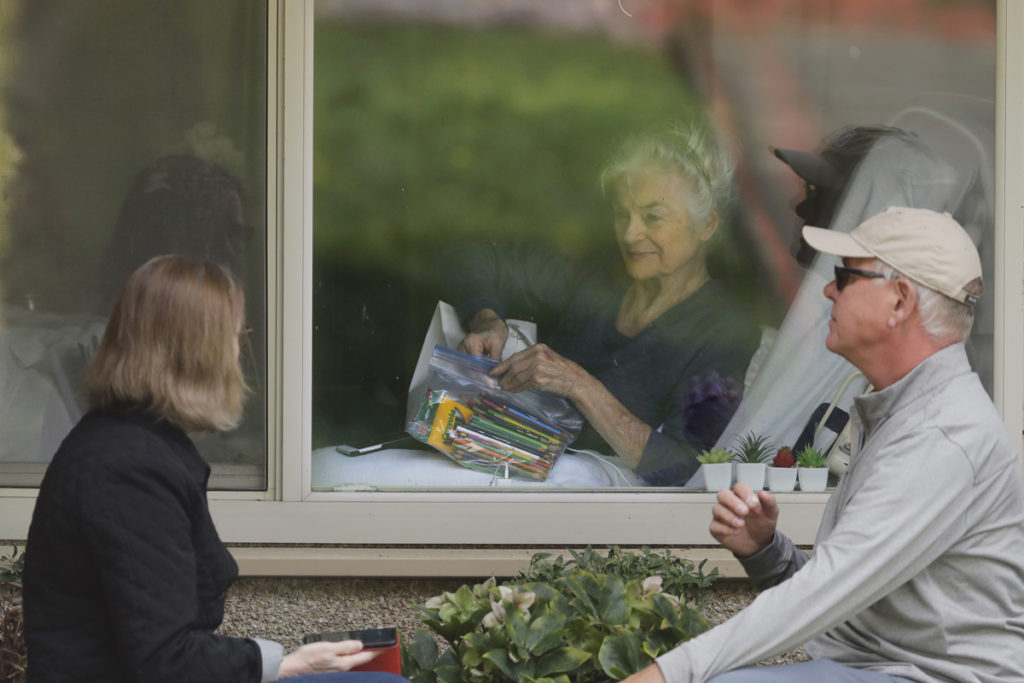 Virus Outbreak US Judie Shape, center, who has tested positive for the new coronavirus, but isn't showing symptoms, opens a care package of art supplies from her daughter and her son-in-law, Tuesday, March 17, 2020, as they talk on the phone and look at each other through a window at the Life Care Center in Kirkland, Wash., near Seattle. (AP Photo/Ted S. Warren)