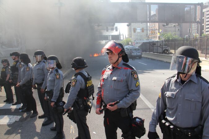 Pennsylvania state troopers keep protesters away from a burning car and vandalized police cars at Broad and Vine streets. (Emma Lee/WHYY)