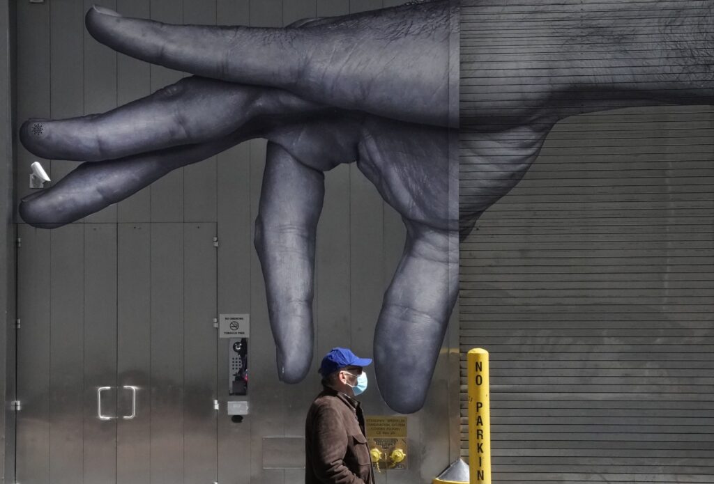 A man in a mask walks past a mural of a hand on the side of a building in N.Y.C in April. A man in a mask walks past a mural of a hand on the side of a building in N.Y.C in April.
