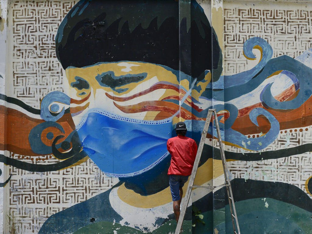 A street artist spray paints a protective face mask over an old mural featuring a Venezuelan Indigenous man, in Caracas, Venezuela, on Saturday. Globally, new daily cases hit an all time high, the World Health Organization reports. A street artist spray paints a protective face mask over an old mural featuring a Venezuelan Indigenous man