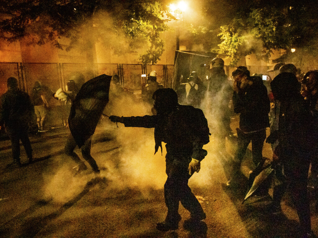 Protesters walk through chemical irritants deployed by federal agents in Portland, Ore. The inspector general of the Justice Department says he is investigating federal officers’ roles in responding to protests in Portland and Washington, D.C. Protesters walk through chemical irritants deployed by federal agents in Portland, Ore. The inspector general of the Justice Department says he is investigating federal officers' roles in responding to protests in Portland and Washington, D.C. (Noah Berger/AP Photo)