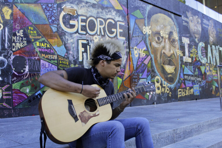 Shane Braswell, who has participated in Black Lives Matter protests this summer, plays guitar in front of a mural covering the entrance to an Apple store, Aug. 27, 2020 in Portland, Ore. Braswell said President Donald Trump's portrayal of Portland as a violent city overcome by mobs of protesters is inaccurate and grossly exaggerated. 