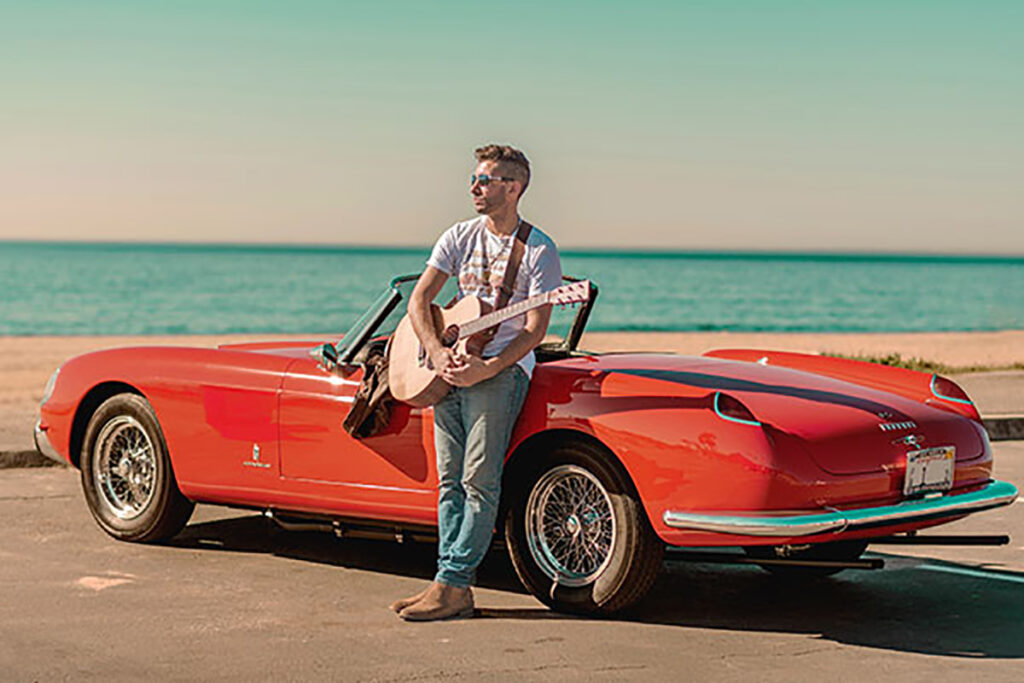 TOM SLESS4x6 Musician Tom Sless with a guitar standing in front of a red