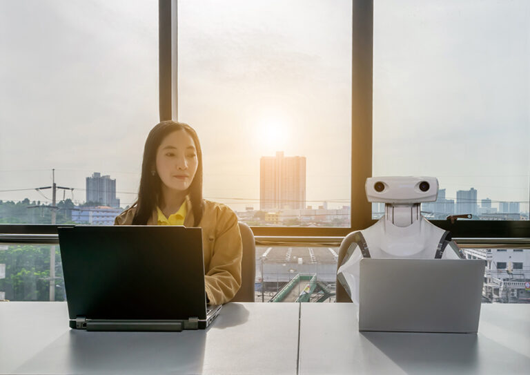 A young woman doing work on a computer, sitting next to a robot working on a computer
