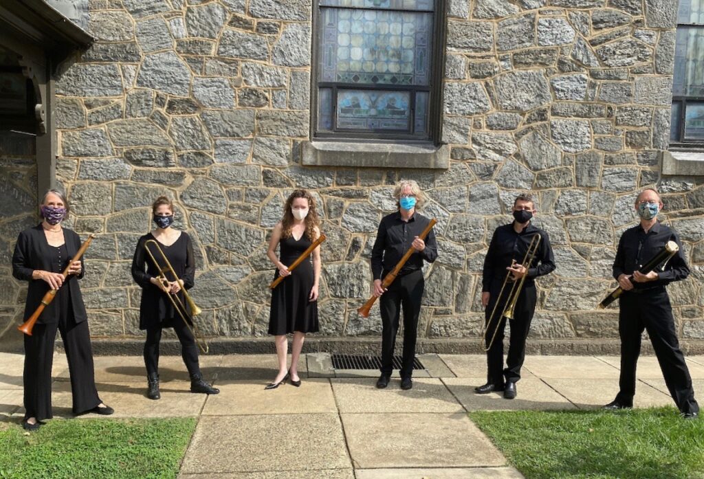Musicians standing outside a stone building holding their instruments and wearing masks