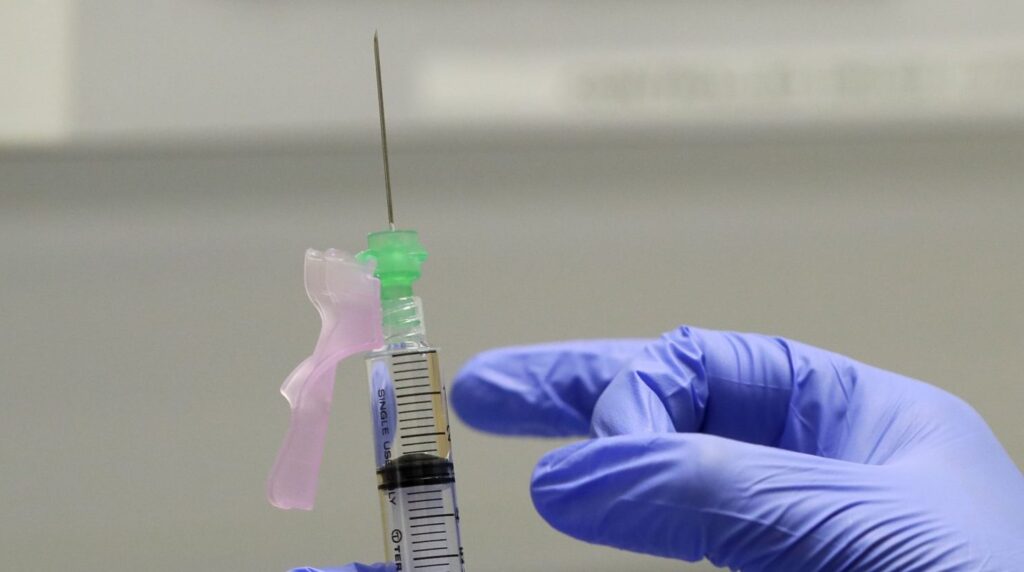 AP20218605422922-e1603237679849-1920×840 A gloved finger prepares the COVID-19 vaccine to administer to a volunteer.
