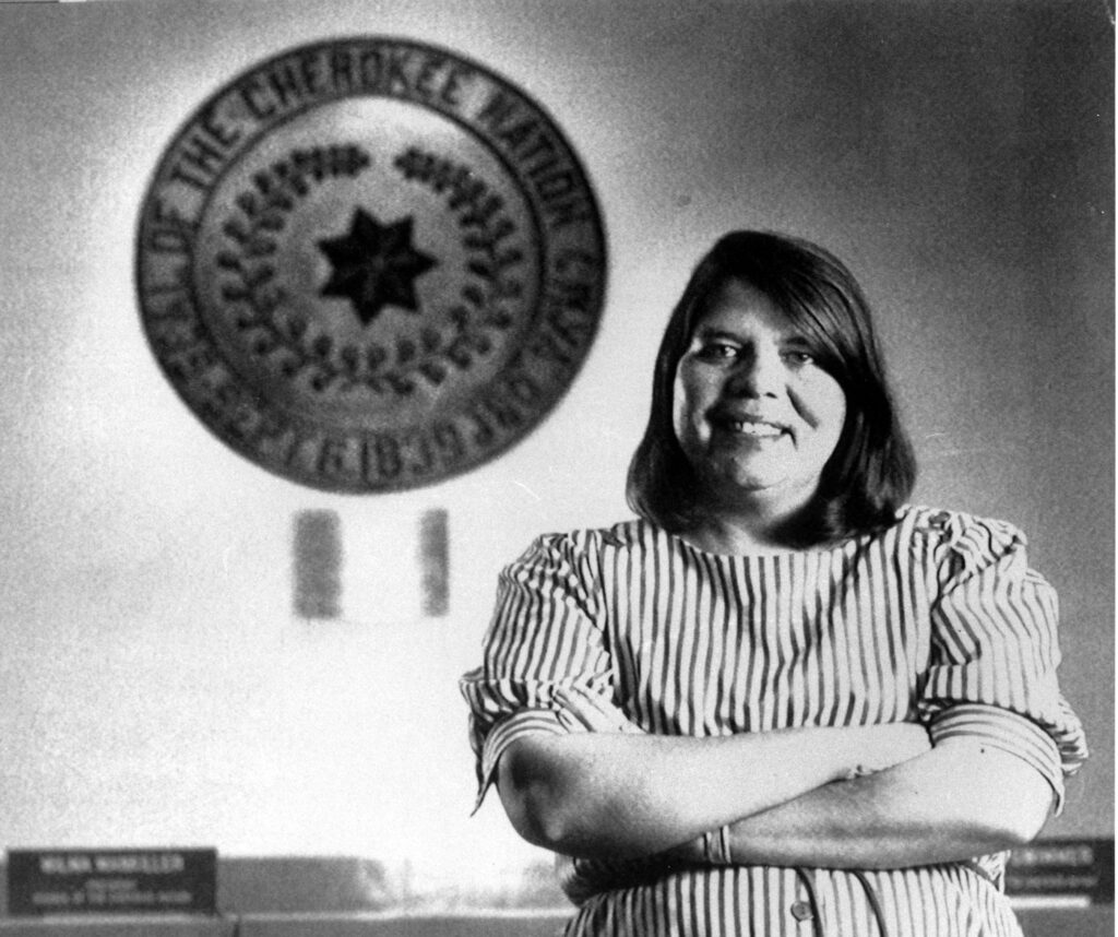WILMA MANKILLER Black and white photography of Wilma Mankiller in front of the tribal emblem at the Cherokee Nation