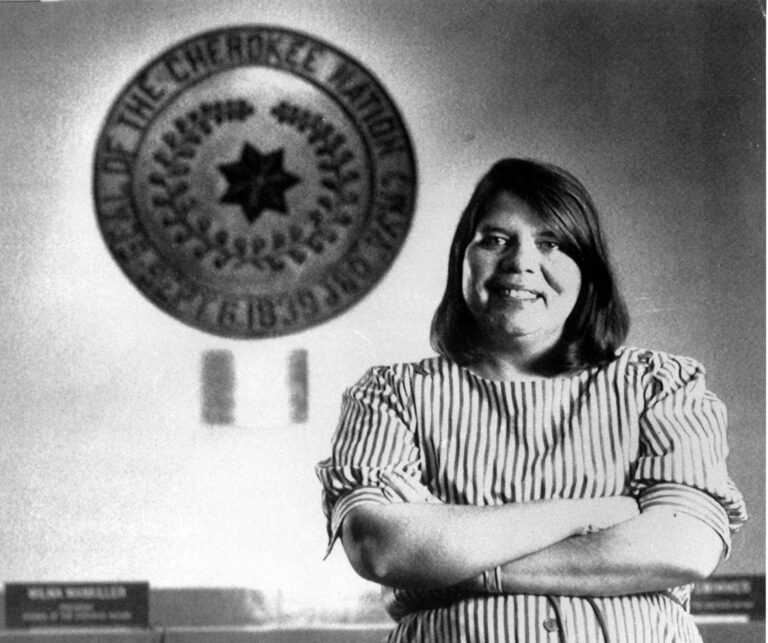 Black and white photography of Wilma Mankiller in front of the tribal emblem at the Cherokee Nation