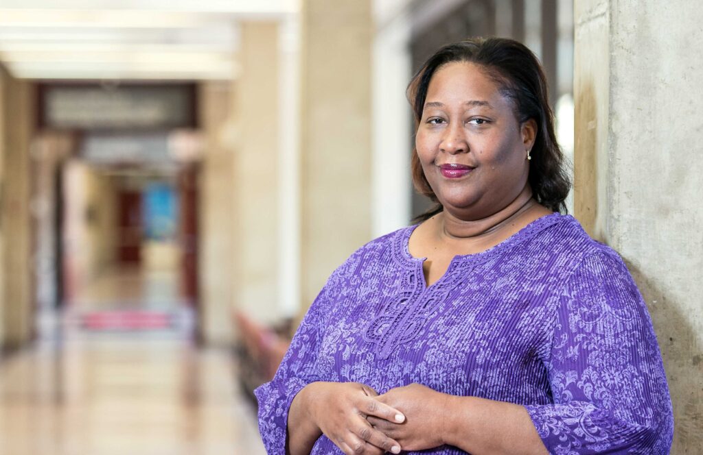 image0-2 Dr. Damali Campbell-Oparaji, a Black woman, wearing a purple shirt stands in a hallway.