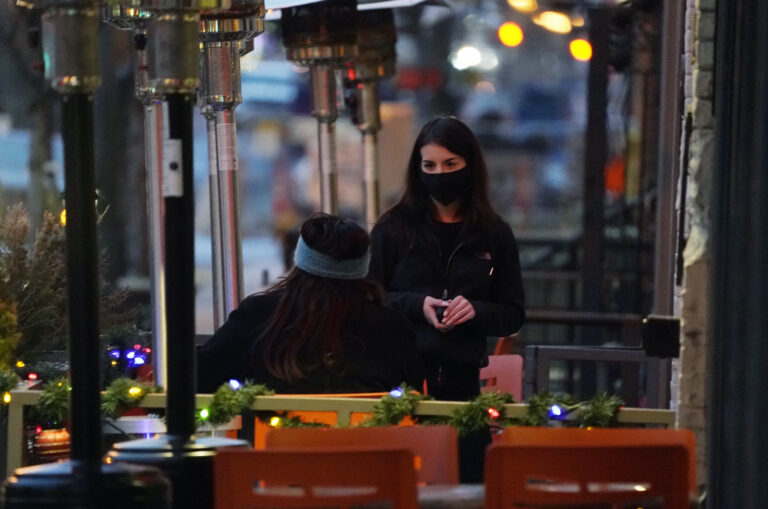 A waitperson wears a face mask while tending to a patron sitting in the outdoor patio of a sushi restaurant late Monday, Dec. 28, 2020, in downtown Denver. (AP Photo/David Zalubowski)