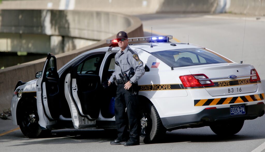 state-police-protests-2018-Pittsburgh-e1592326374868 A Pennsylvania State Trooper stands beside his vehicle parked on the entrance ramp to Interstate 376 out of Pittsburgh as protesters block an intersection in downtown Pittsburgh, on Tuesday, June 26, 2018. (Keith Srakocic/AP Photo)