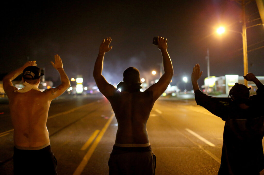 Outrage In Missouri Town After Police Shooting Of 18-Yr-Old Man Demonstrators raise their arms and chant,