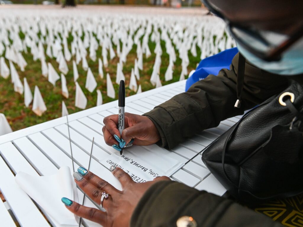 Patrice Howard writes on white flags before planting them to remember her recently deceased father and close friends in November at “IN AMERICA How Could This Happen…,” a public art installation in Washington, D.C. Led by artist Suzanne Firstenberg, volunteers planted white flags in a field to symbolize each life lost to COVID-19 in the U.S. Patrice Howard writes on white flags before planting them to remember her recently deceased father and close friends