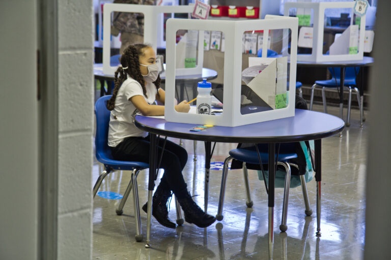 A student, wearing a face mask, sits at an isolated desk at school