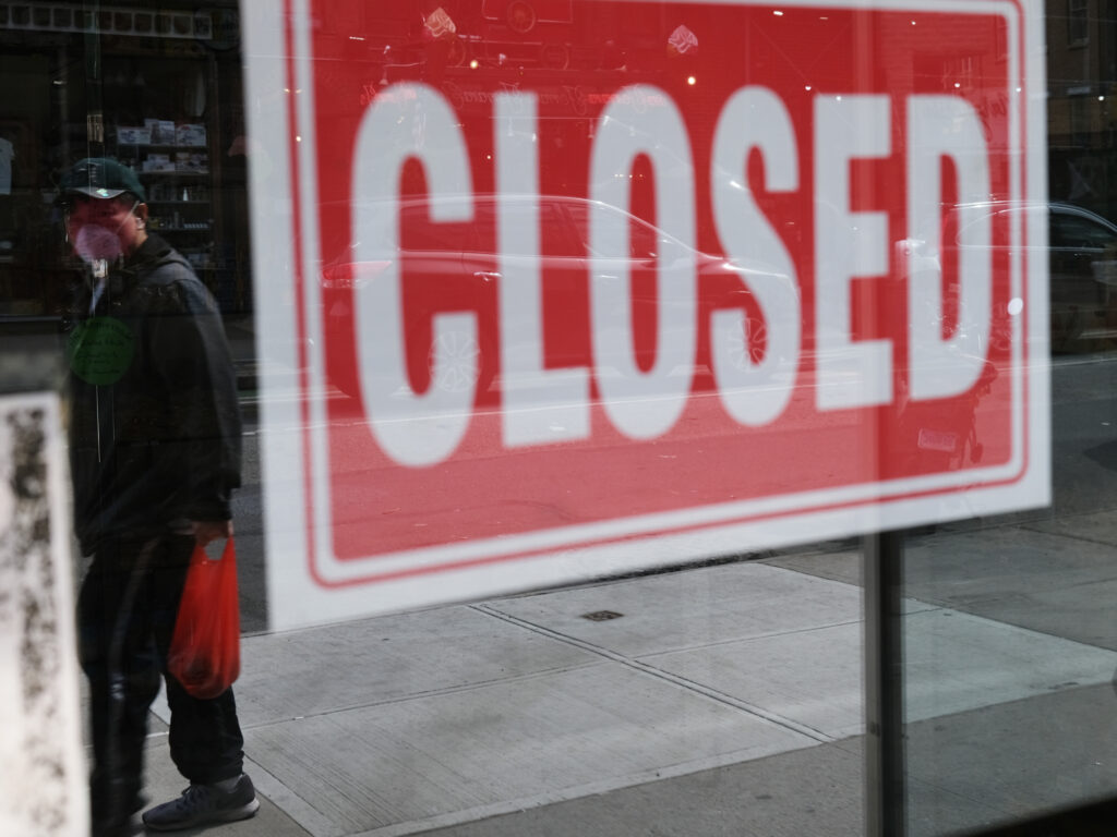 A store in New York City stands closed on April 21, 2020. A year after the pandemic started to spread, millions of Americans are still unemployed. A person's reflection is seen in a store window that has a closed sign affixed to it