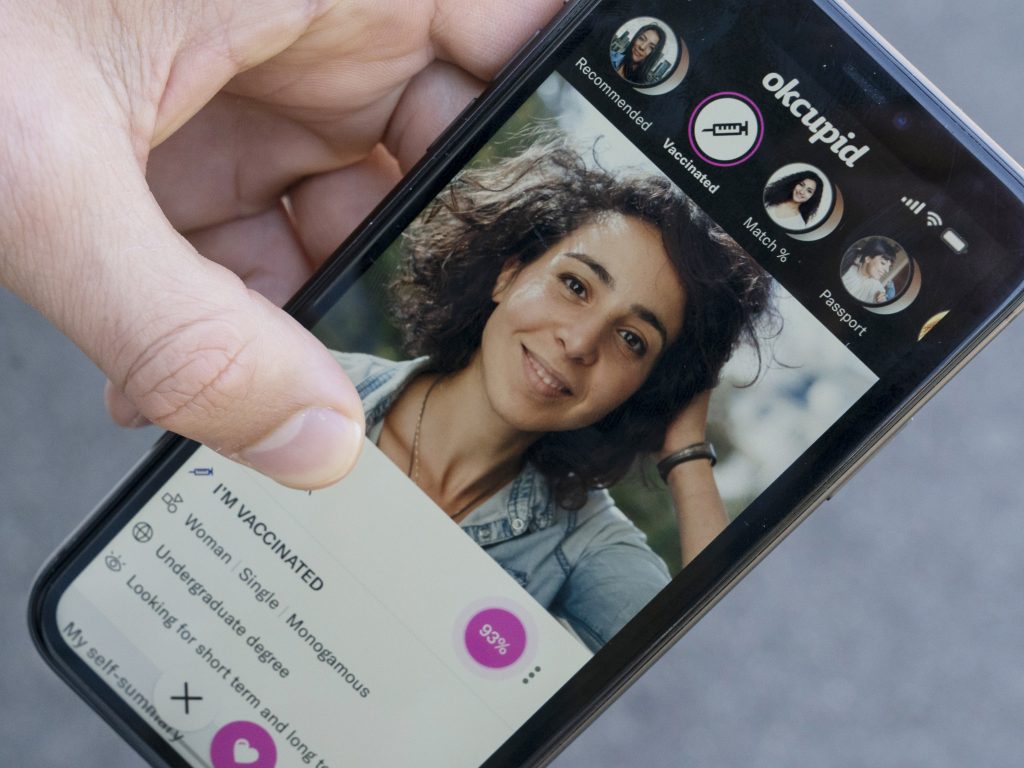 A sample page on the OkCupid app is held for a photograph showing the “I’m vaccinated” checkmark. A sample page on the OkCupid app is held for a photograph showing the