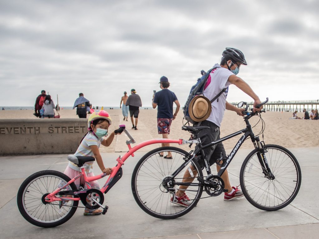 A girl and her father wear face masks while they push their bikes last summer in Hermosa Beach in the Los Angeles area. There aren’t yet coronavirus vaccines approved in the U.S. for kids under 12 — which means they should keep masking, according to the CDC. A girl and her father wear face masks while they push their bikes in Hermosa Beach
