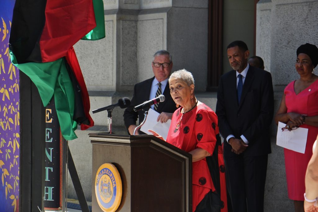 Helen Salahuddin speaks from a podium, with a Black Liberation Flag visible in the background
