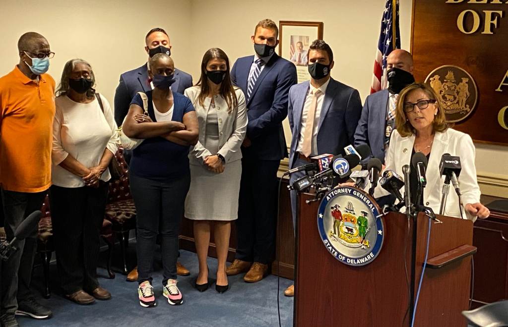 Gang indictment Delaware Attorney General Kathleen Jennings speaks from a podium as victims' family members stand to her left