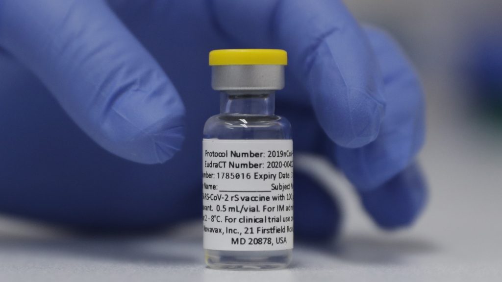 A vial of the experimental Novavax coronavirus vaccine is ready for use in a London study in 2020. Novavax’s vaccine candidate contains a noninfectious bit of the virus — the spike protein — with a substance called an adjuvant added that helps the body generate a strong immune response. A gloved hand is seen about to pick up a vial of the experimental Novavax COVID-19 vaccine