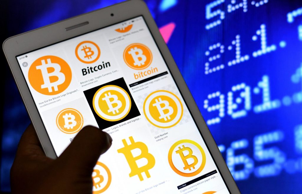 In this photo illustration, Bitcoin logos seen displayed on A hand holds up a phone with Bitcoin logos on it, against the backdrop of a screen with numbers on it