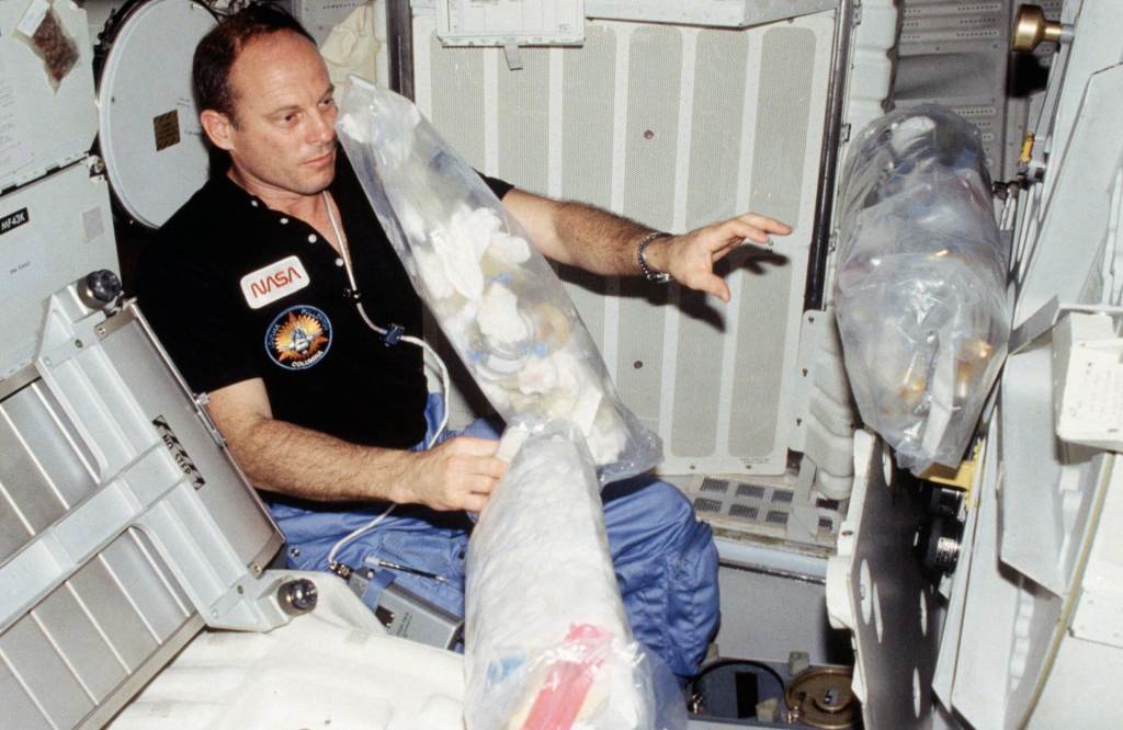 Astronaut Jack R. Lousma, STS-3 commander, wearing communication kit assembly mini headset, gathers three freefloating plastic trash bags filled with empty containers, paper towels, straws, etc. Lousma will stow them in a designated stowage volume. (NASA)