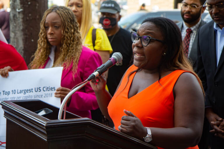 Kendra Brooks speaks from a podium at a press conference, with elected officials behind her