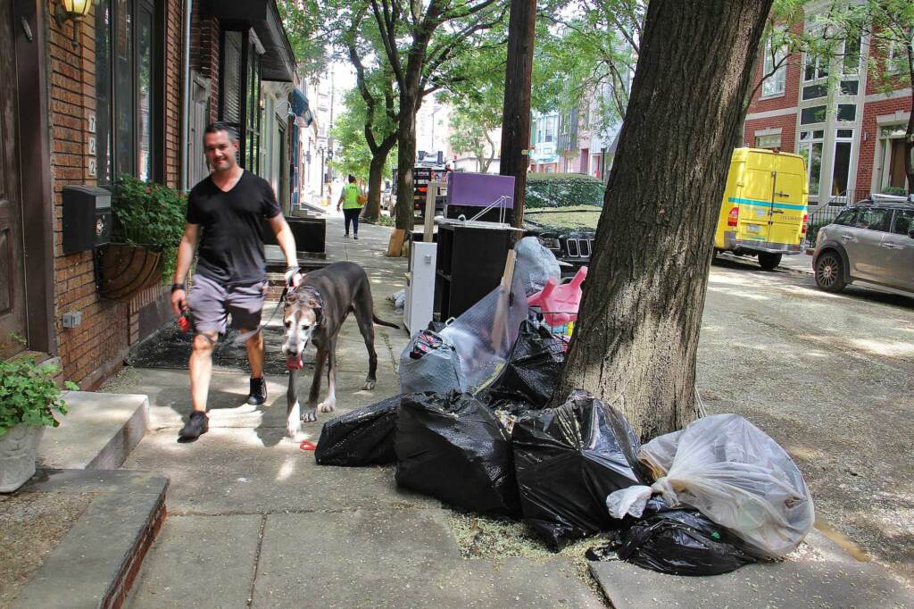Water damaged possessions are piled at the curb on 6th Street as a resident and their dog walk by