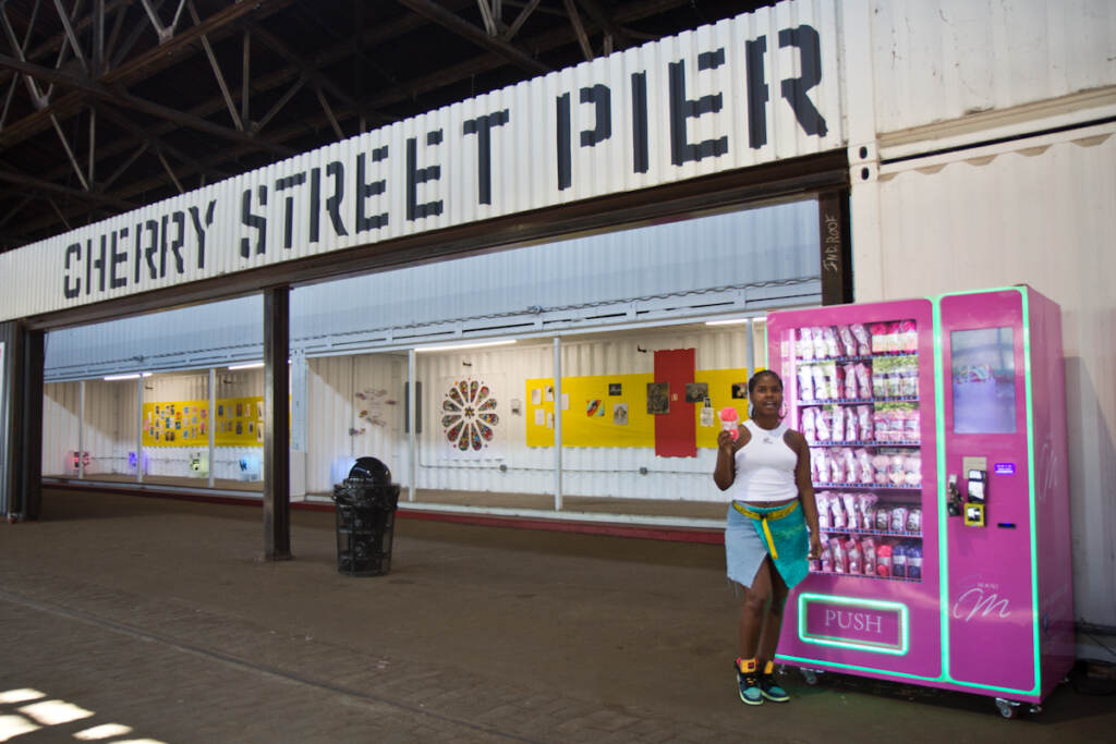 Emani Outterbridge holds up yarn in front of one of her vending machines, with Cherry Street Pier signage behind her