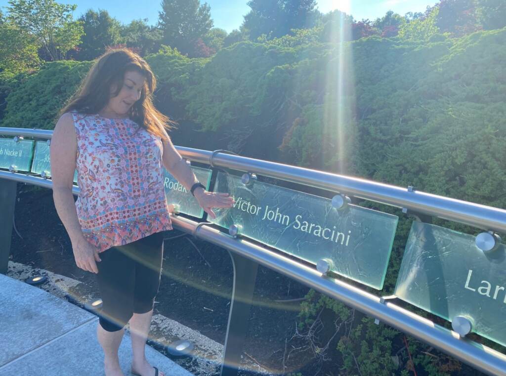 FEATURED-IMAGE-scaled-e1630605594324-1920×1426 Ellen Saracini touches a memorial plaque for her husband Victor that is illuminated by late afternoon sunlight. Victor, who piloted one of the doomed planes before being killed by terrorists, is one of 2,973 victims of the terror attacks remembered at the Garden of Reflection. (Brett Sholtis / WITF)