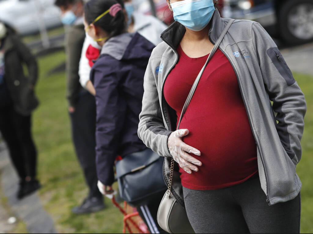 A pregnant woman wearing a face mask and gloves holds her belly as she waits in line for groceries at St. Mary’s Church in Waltham, Mass., in May. The Centers for Disease Control and Prevention on Wednesday again urged all pregnant women to get the COVID-19 vaccine. A pregnant woman wearing a face mask and gloves holds her belly as she waits in line for groceries