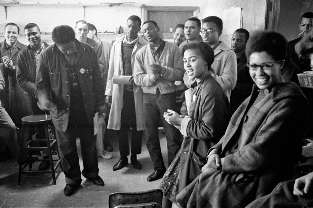 SNCCPhoto1_Richardson rev James Forman leads singing in the SNCC office on Raymond Street in Atlanta, (from left) Mike Sayer, MacArthur Cotton, Forman, Marion Barry, Lester McKinney, Mike Thelwell, Lawrence Guyot, Judy Richardson, John Lewis, Jean Wheeler, and Julian Bond, 1963, Danny Lyon