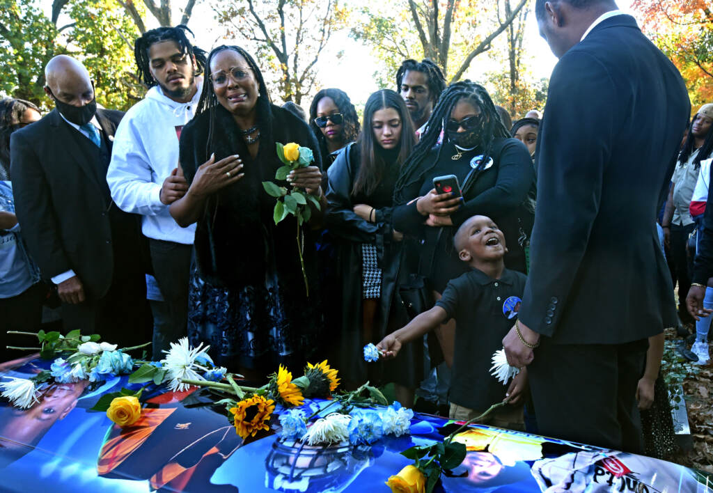 2021-11-08-a-saul-jassan-funeral On Nov. 8, at the Sunset Memorial Park Cemetery in Pennsauken, Jassan Lindsey's paternal grandmother, Alma Lindsey, talks about how much her grandson meant to her at his casket; at right, Jassan's younger brother, Kason Lindsey, 4 — too young to comprehend the loss — holds a flower. (April Saul for WHYY)
