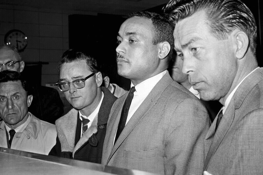 Thomas 15X Johnson Khalil Islam, center, is booked as the third suspect in the slaying of Malcolm X, in New York, March 3, 1965. Islam, previously known as Thomas 15X Johnson, one of two men convicted in the assassination of Malcolm X, is set to be cleared after more than half a century, with prosecutors now saying authorities withheld evidence in the civil rights leader's killing, according to a news report Wednesday, Nov. 17, 2021. Detective John Keeley is at right. (AP Photo, File)
