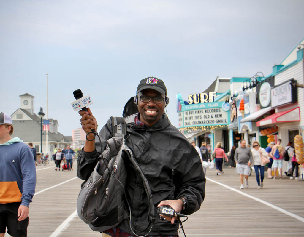 Kenny Burns Newsroom Staff Cover Photo A reporter poses on a beach boardwalk with his recording equipment, smiling and holding his microphone aloft.