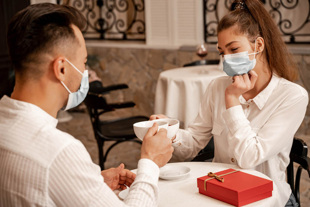 YOK-1-21-bigstock-Young-Couple-In-Medical-Masks–422998292 Young couple in medical masks clinking cups of tea near gift box on table in cafe