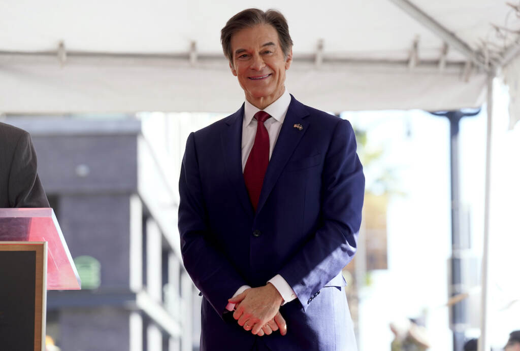 Mehmet Oz Mehmet Oz stands with hands clasped in front of him, wearing a dark blue suit and a red tie.