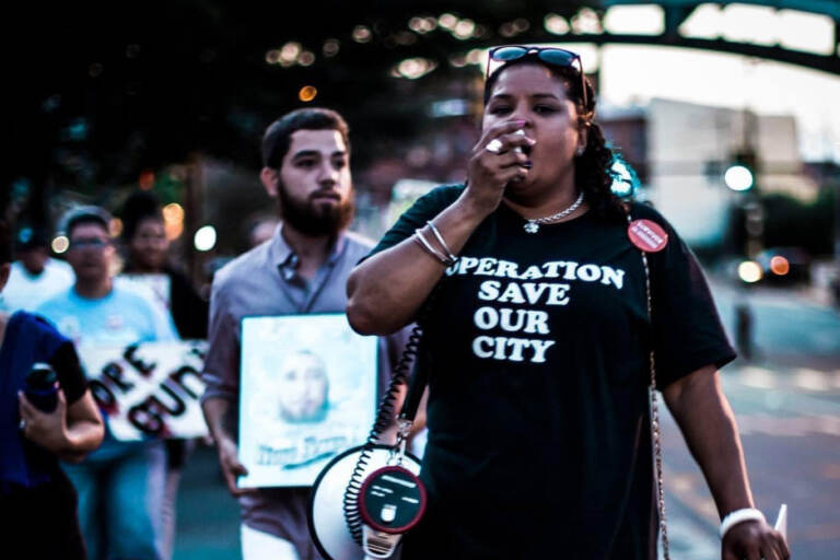 Rosalind Pichardo leading a crowd of people with a megaphone, wearing an 
