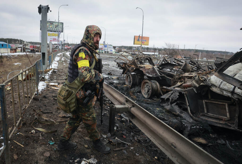 APTOPIX Russia Ukraine War An armed man stands by the remains of a Russian military vehicle in Bucha, close to the capital Kyiv, Ukraine, Tuesday, March 1, 2022. Russia on Tuesday stepped up shelling of Kharkiv, Ukraine's second-largest city, pounding civilian targets there. Casualties mounted and reports emerged that more than 70 Ukrainian soldiers were killed after Russian artillery recently hit a military base in Okhtyrka, a city between Kharkiv and Kyiv, the capital.