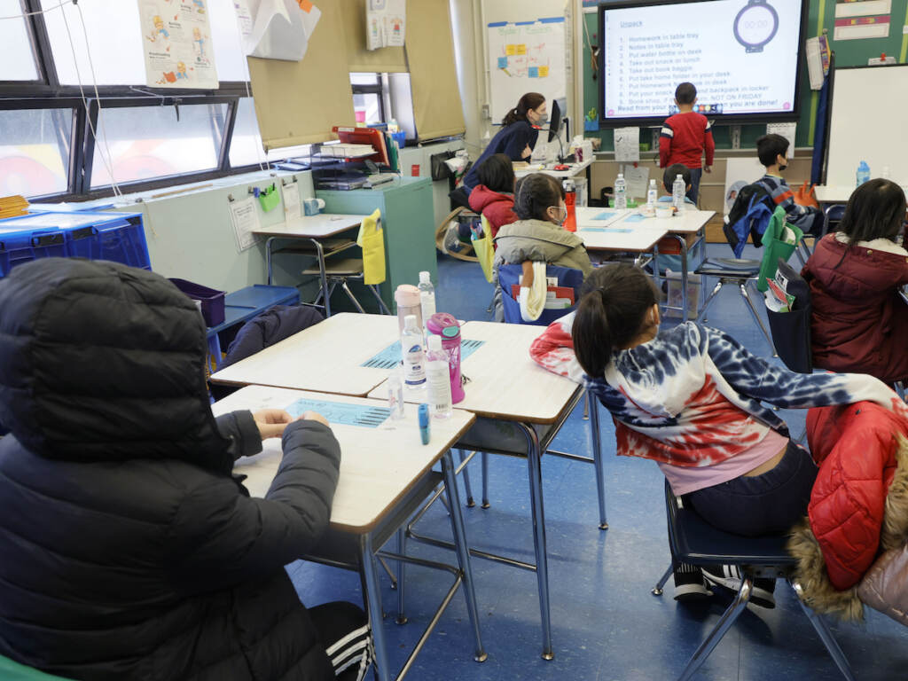 Open windows and portable air cleaners can help improve ventilation, but many schools in the U.S. are more than 45 years old and need a complete overhaul of their HVAC systems. Seated students are shown from behind, seated at desks, looking towards a teacher and student at the front of the classroom.