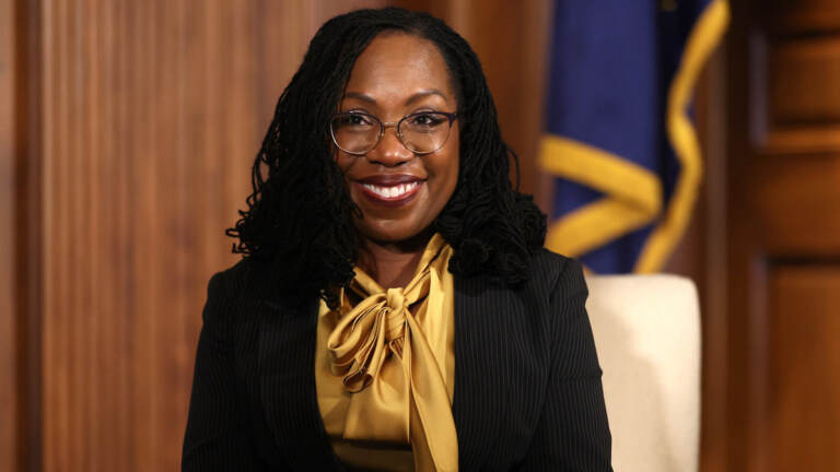 A portrait-style photo of U.S. Supreme Court nominee, Ketanji Brown Jackson. She's smiling, wearing a gold-colored blouse and black jacket.