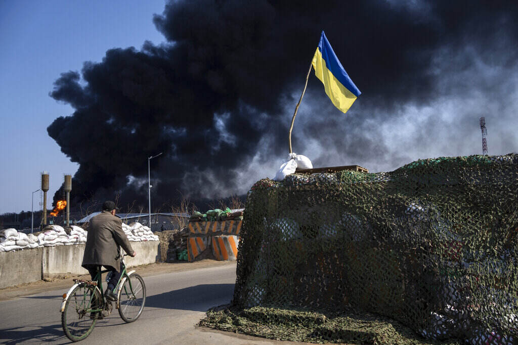 APTOPIX Russia Ukraine War A man rides a bicycle past a structure with the Ukrainian flag flying on top, as smoke billows in the background.