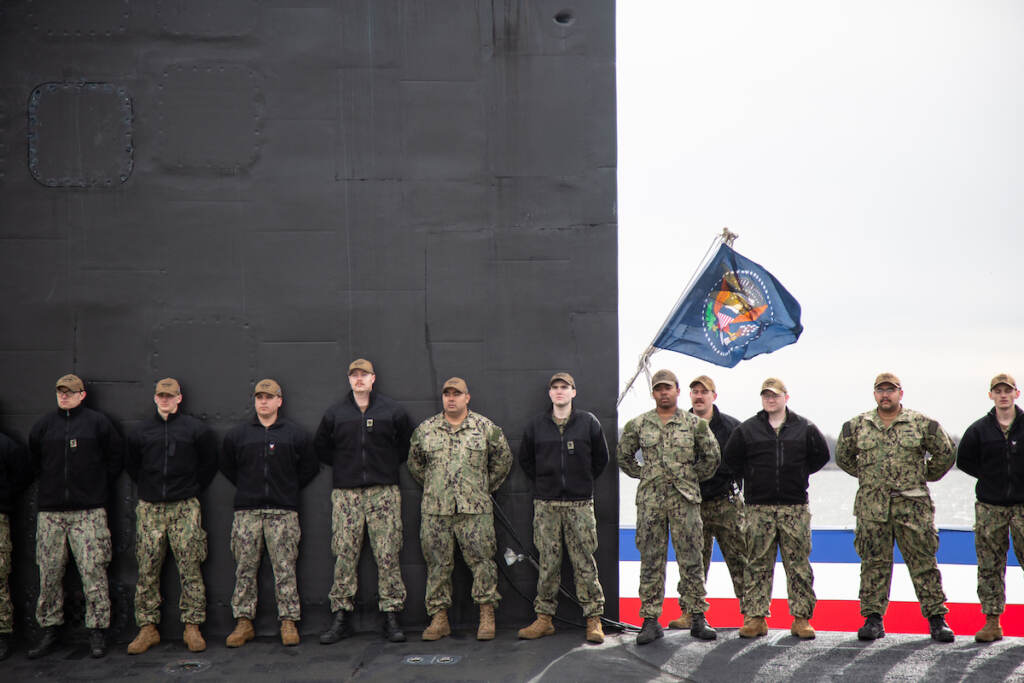 2022 04 01 – e cohen- dress rehearsal for submarine commission- Delaware Waterfront-2654 The crew of the USS Delaware stand in front of a flag of the Presidental Seal while on their submarine during the dress rehearsal for the official commission ceremony for the new submarine.