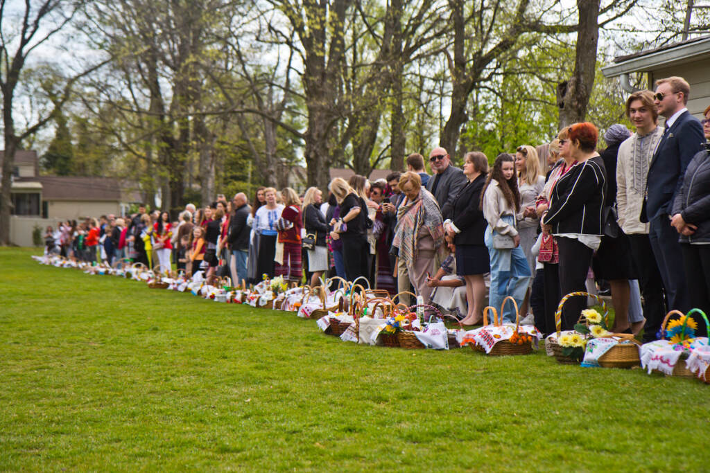 2022 04 25 k paynter ukrainain easter services-4 A row of Ukrainian American parishioners stand on the edge of a green field, with Easter baskets in front of them.